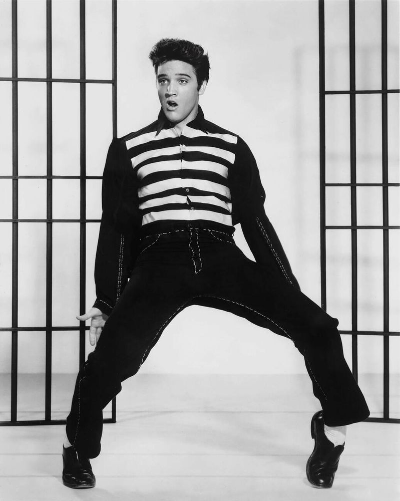 Singer and actor Elvis Presley — in costume, dressed in a black-and-white striped shirt and matching black denim jacket and jeans — captured facing the camera while standing in front of a set of open prison cell doors, his knees bent, left foot on tiptoes, arms by his side as he looks off-camera with his mouth open, mid-thrust, in a full-length black-and-white shot for the film Jailhouse Rock (1957)