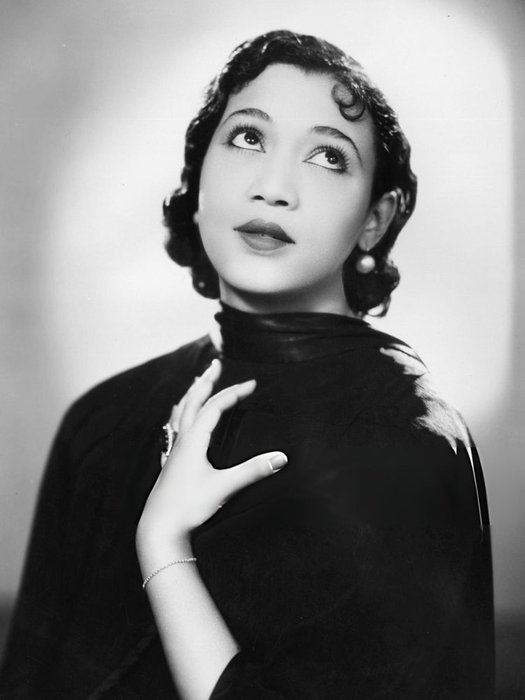 Actress and singer Elisabeth Welch — dressed in a black gown and wrapped in a black cape — captured almost in profile while seated and posed against a well-lit background, looking upwards, her left hand resting on her chest, in a medium-length black-and-white studio portrait taken by the photographer Sasha