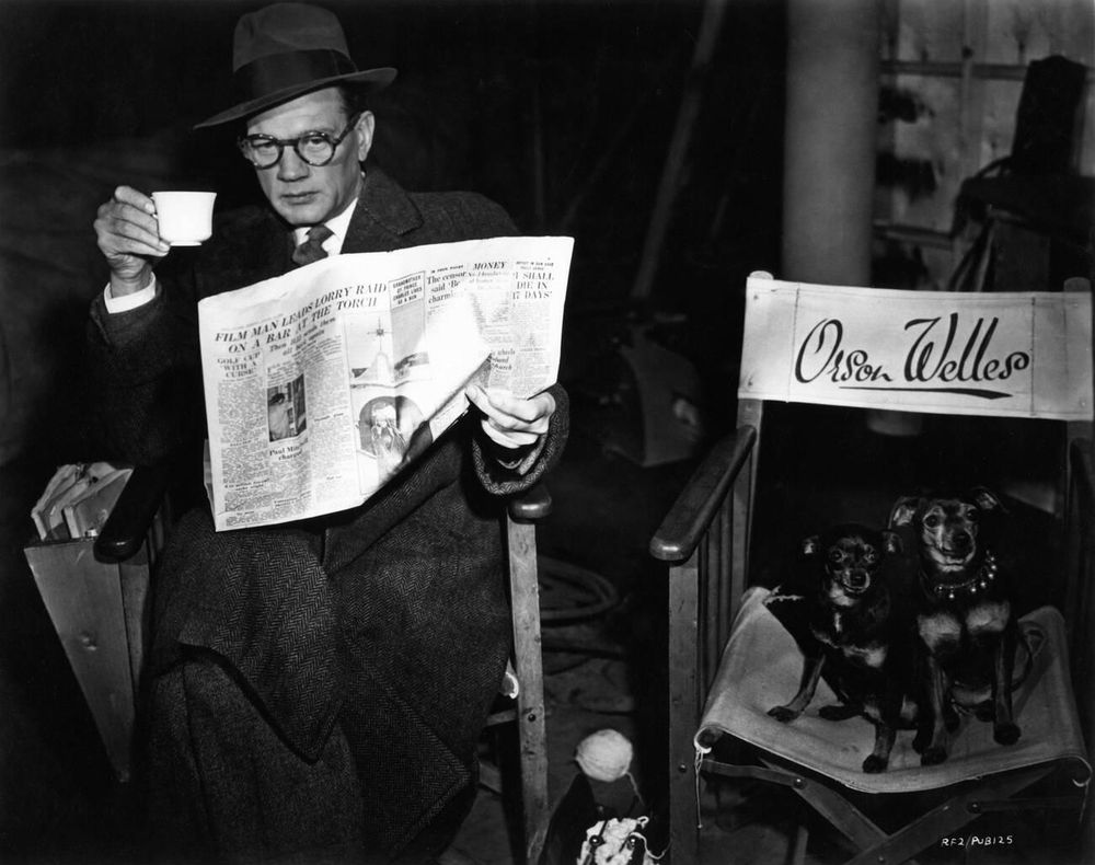 Actor Joseph Cotten — in costume, dressed in a dark-coloured suit, tie, overcoat and fedora hat — captured facing the camera while sitting on a director's chair, clutching a newspaper in his left hand and a white tea cup in his right, a director's chair inscribed with the name "Orson Welles" positioned beside him, upon which two Miniature Pinscher dogs sit, in a medium-length shot taken during a break from filming The Third Man (1949)