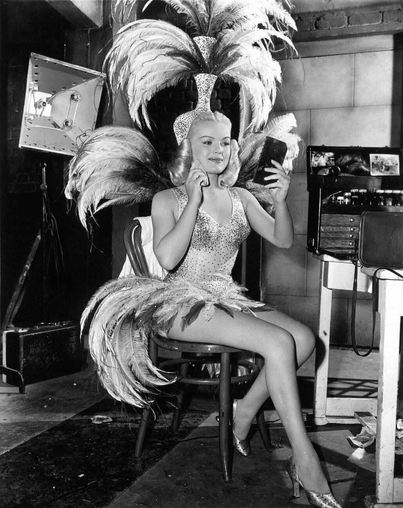 Actress, singer and dancer June Haver — in costume, dressed in a light-coloured sparkling sequined showgirl ensemble with dramatic feathered headdress and accoutrements — captured sitting almost in profile while holding a hand mirror in front of her face and dabbing a powder puff against her right cheek, a large make-up kit positioned to her left, in a full-length black-and-white shot taken during a break from filming on the set of The Dolly Sisters (1945)