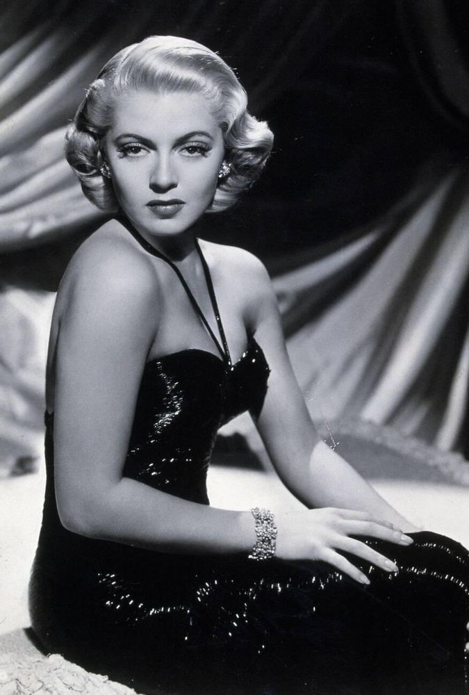Actress Lana Turner — dressed in a sparkling black strapless gown with thin halterneck — captured sitting in profile in plush surroundings, face turned to the camera, looking directly into the lens, her hands resting on her legs in a medium-length black-and-white shot for Slightly Dangerous (1943)
