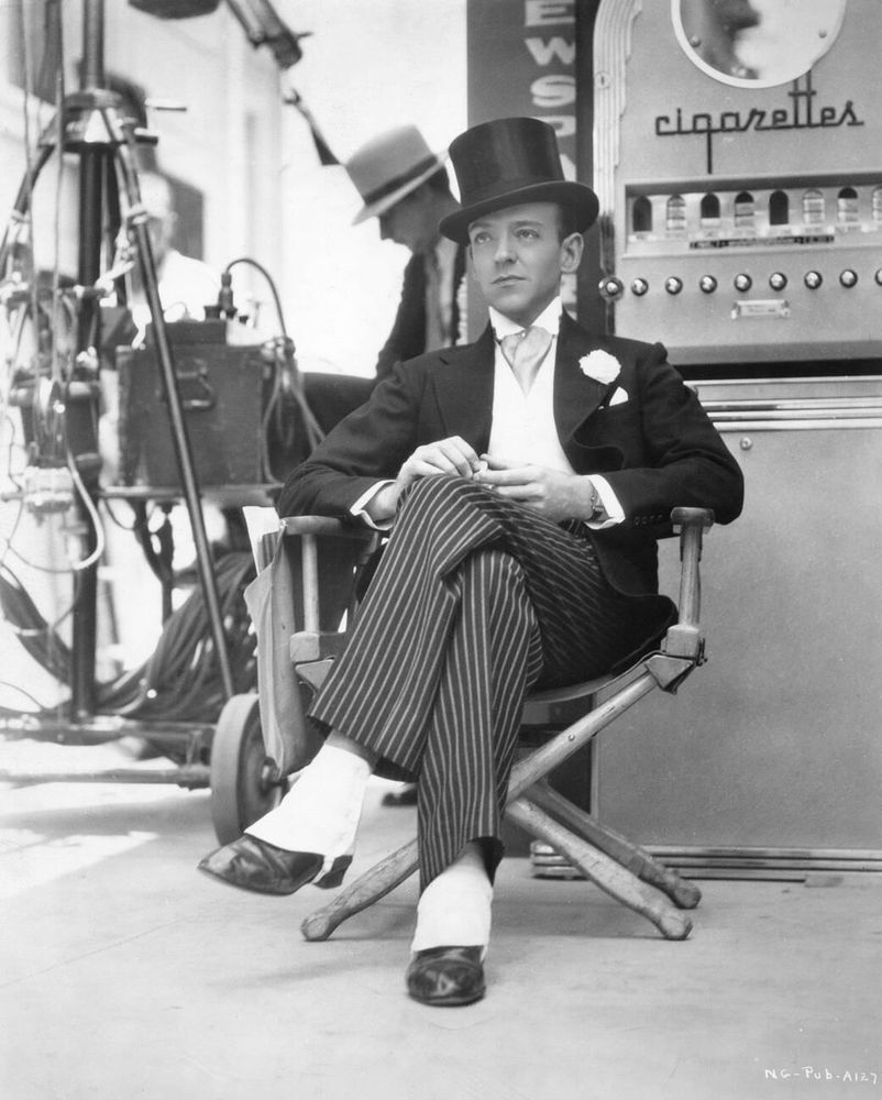 Actor and dancer Fred Astaire — dressed in white tie and tails, black pinstripe trousers and wearing a black top hat — captured facing the camera while sitting on a director's chair, looking off-camera, holding a small white object in his hands, a large cigarette vending machine prop behind him alongside a crew member and filming equipment, in a full-length black-and-white shot taken during a break from filming on the set of Swing Time (1936)
