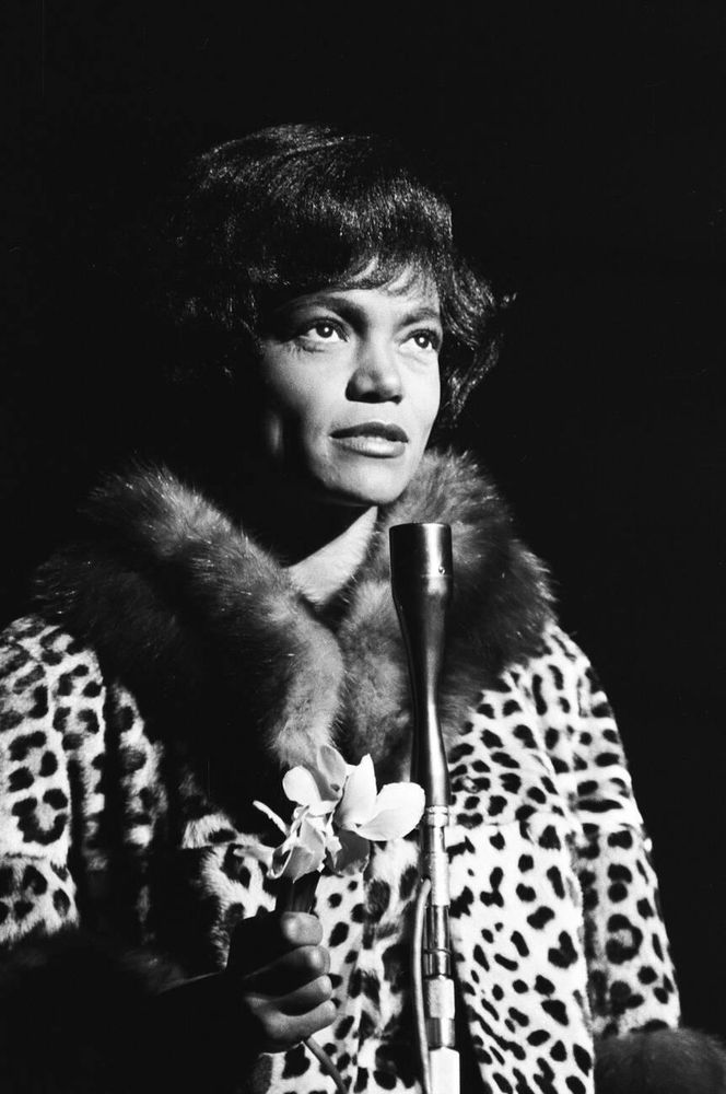 Singer and actress Eartha Kitt — dressed in a leopard print coat with dark-coloured fur-trimmed collar and cuffs — captured facing the camera while standing in front of a microphone, looking upwards, a white flower held in her right hand, in a medium-length black-and-white shot taken during a performance at The Talk of the Town in London