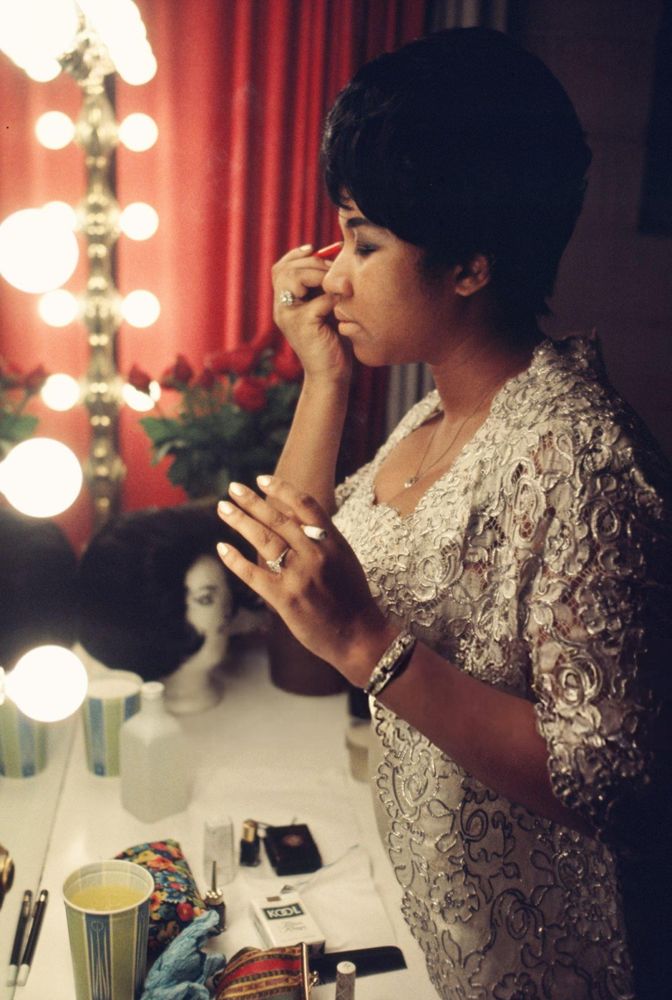 Musician Aretha Franklin — dressed in a gold gown with textured patterned layers — captured in profile, standing before a lighted vanity mirror, eyes closed as she uses her right hand to apply eyeliner to her right eye, a lit cigarette in her left hand, make-up items, a packet of cigarettes and a cup of liquid on the table in front of her, a wig on a head mannequin and a vase of red roses nearby, in a medium-length colour shot taken by Walter Iooss in Franklin's dressing room, backstage at the Symphony Hall in Newark, New Jersey