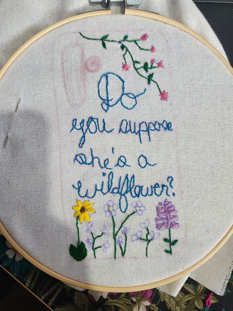 Embroidery loop with partially complete project.
Outline of cellphone back.
Words embroidered in blue "do you suppose she's a wildflower?"
7 different flower doodles at various points of completion.