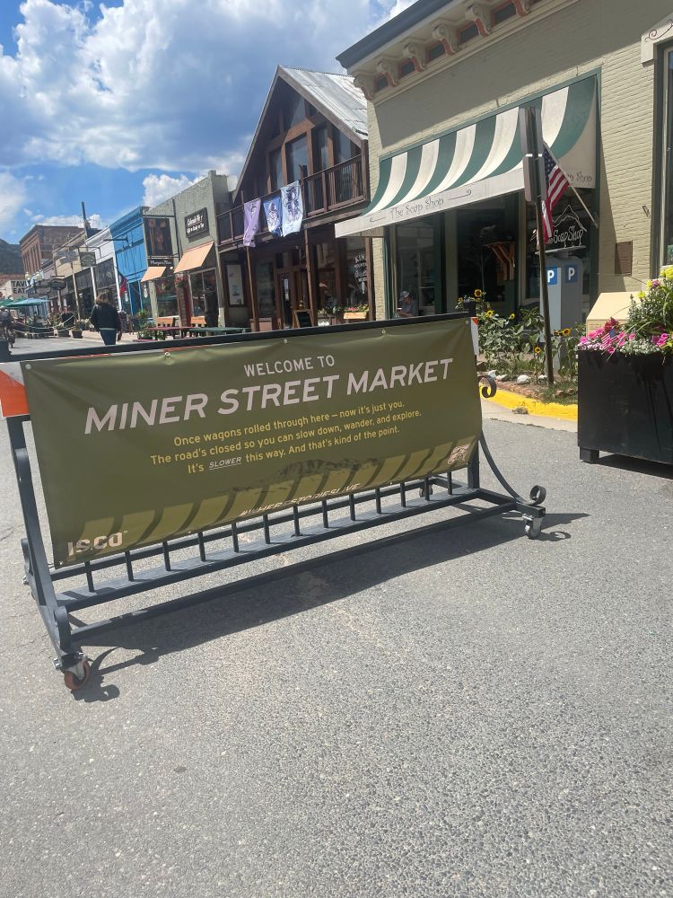 Sign showing Miner Street opened for people to walk. It says Miner Street Market. 

The smaller text reads: Once Wagons rolled through here - now it is just you. The road is closed so you can so slow down, wander and explore. It is SLOWER and that is kinda the point. 

The sign is flanked by flower boxes.