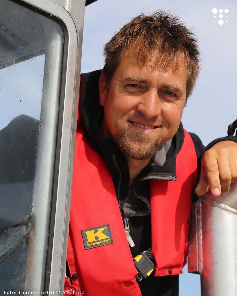 Ein Mann mit roter Schwimmweste lächelt in die Kamera und stützt sich auf einem silbernen Geländer ab.