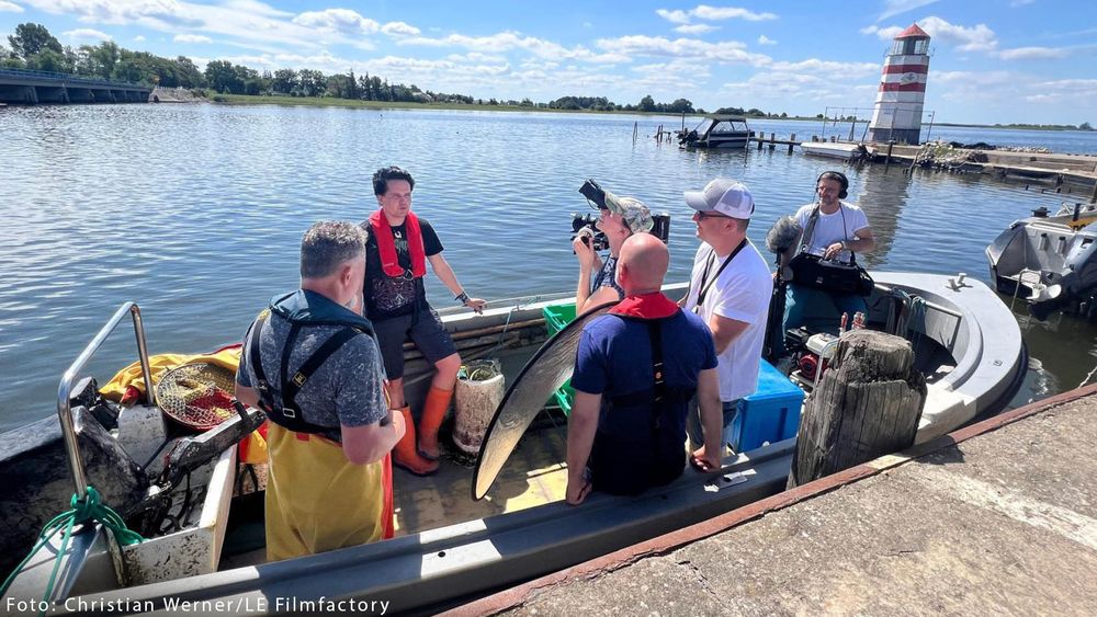 Eine Gruppe von fünf Personen befindet sich auf einem kleinen Boot auf einem ruhigen Gewässer bei sonnigem Wetter. Eine Person mit roter Schwimmweste sitzt auf dem Bootsrand und wird interviewt, während eine weitere Person mit einer Kamera das Geschehen filmt. Zwei andere stehen mit einem Reflektor und anderem Equipment in der Nähe und scheinen zur Filmcrew zu gehören. Ein Tonassistent mit Mikrofon steht rechts. Im Hintergrund sind rechts ein rot-weißer Leuchtturm sowie ein Steg und ein Boot zu sehen. Das Wasser spiegelt den blauen Himmel wider. Am unteren Bildrand steht der Bildnachweis: „Foto: Christian Werner/LE Filmfactory“.