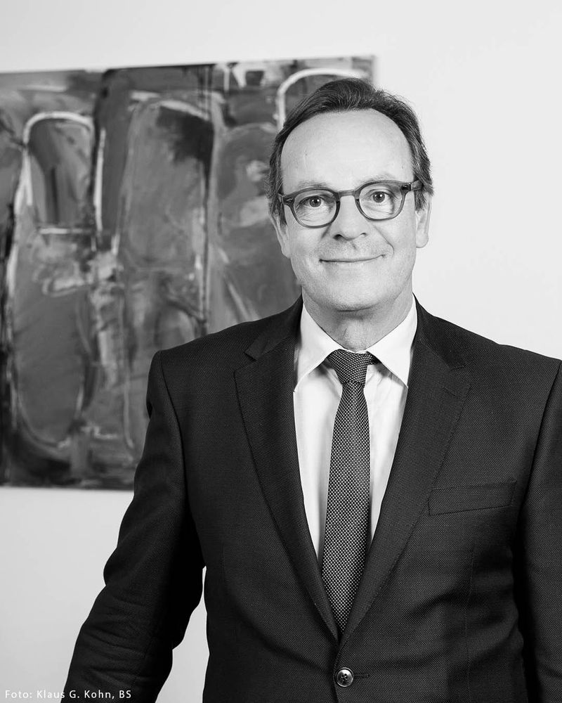Schwarz-Weiß-Porträt von Folkhard Isermeyer in einem formellen Anzug mit Krawatte. Er trägt eine Brille mit dunklem Rahmen und hat kurzes Haar. Er lächelt dezent in die Kamera und hat eine professionelle, selbstbewusste und freundliche Ausstrahlung.
Im Hintergrund ist ein abstraktes Kunstwerk zu sehen, das aus dunklen und hellen Flächen besteht. In der unteren linken Ecke befindet sich der Hinweis „Foto: Klaus G. Kohn, BS