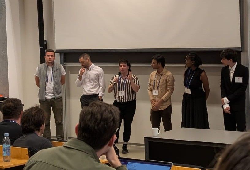 Cedar answers an audience question, alongside 5 other speakers, as audience members look on