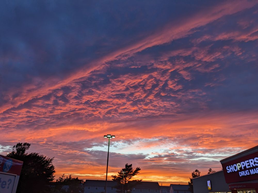 A sunset against the clouds. The clouds have a strange Vee like shape, and are a mixture of dark blues, violet greys, and pink gold with orange as we go from the upper left to the lower right. The sun is technically sunk under the edge of the horizon, leaving dark silhouettes of trees and houses in the foreground.