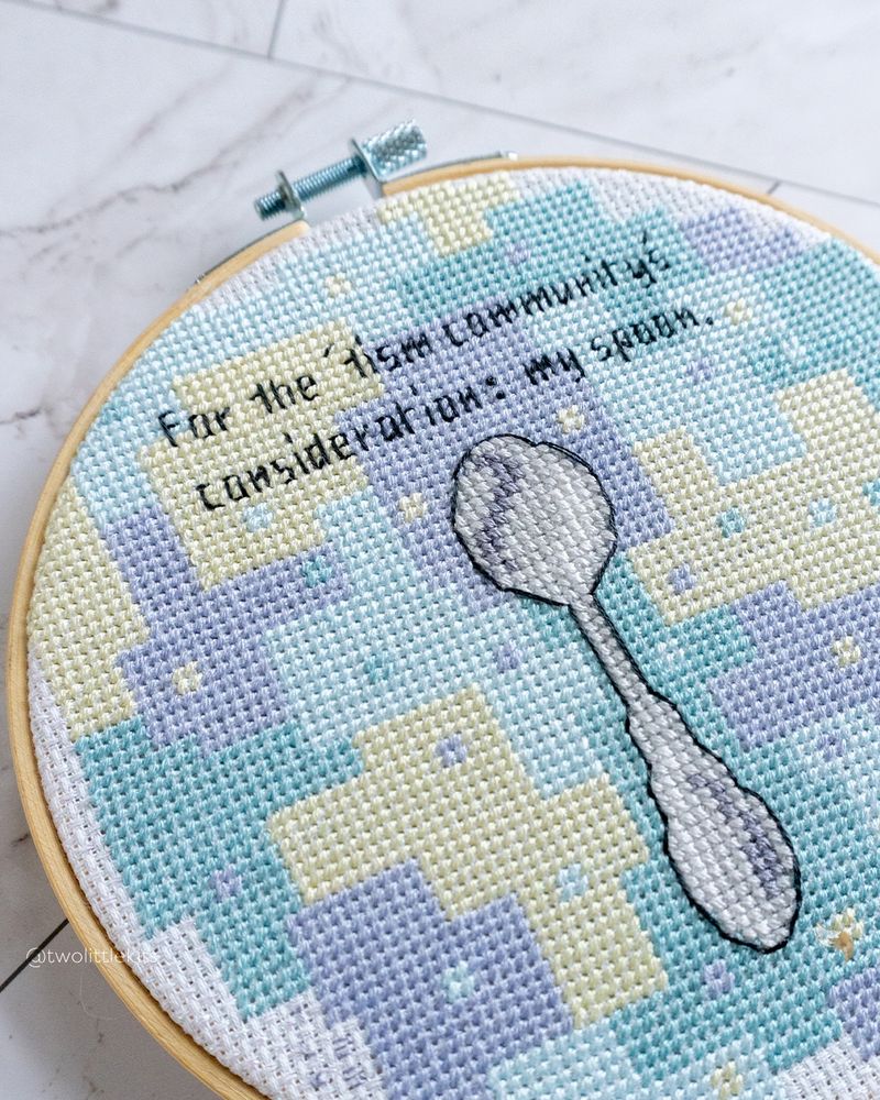 A close-up photo of a cross-stitch piece in a 6" embroidery hoop. It's mostly blues and greens in a geometric/blocky style, but in the middle it has a silver spoon. Above the spoon it reads "For the 'tism community's consideration: my spoon."