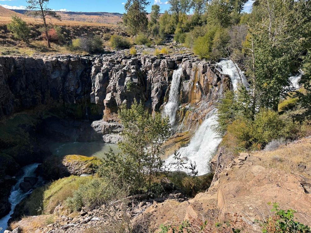 A scenic waterfall cascading over rocky cliffs surrounded by lush greenery and trees under a clear blue sky.