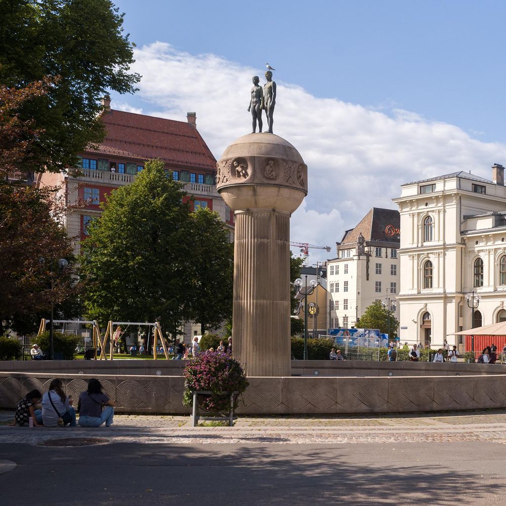 A tall column with sculptures of two figures on top, located in a city square. A seagull sits atop the sculpture. Surrounding buildings and trees are visible, along with people sitting and walking nearby.