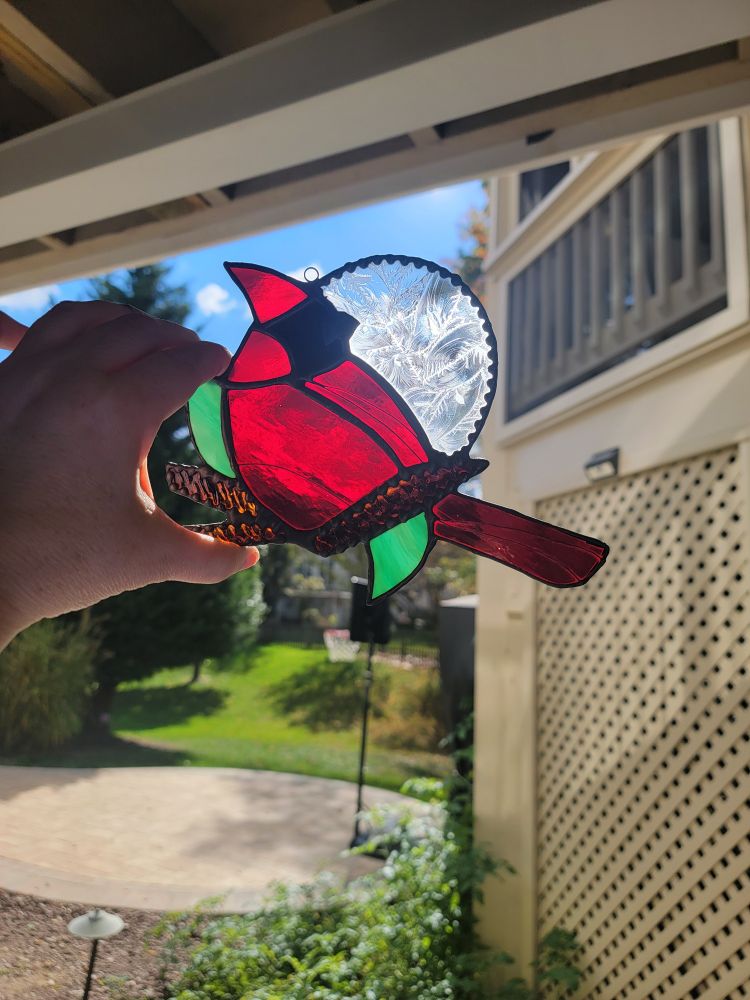 A suncatcher being held up to the sunlight. It is a cardinal sitting on a branch with green leaves.