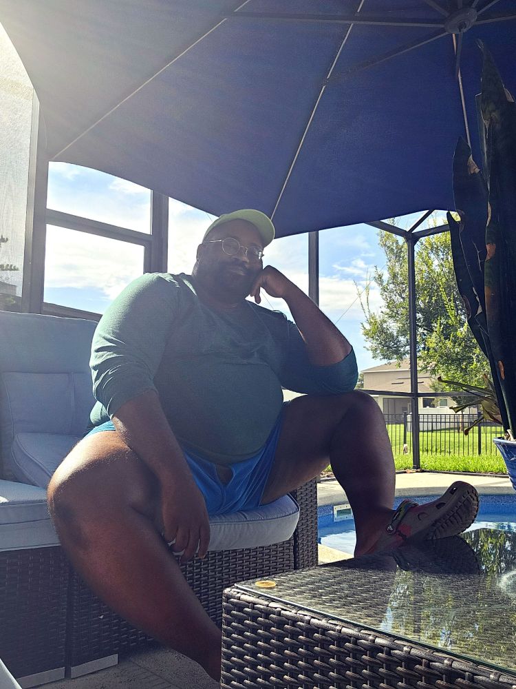 Sitting outside on the back patio, wearing a green shirt, blue undies  a green hat, and brown crocs. Sun is flowing in behind me