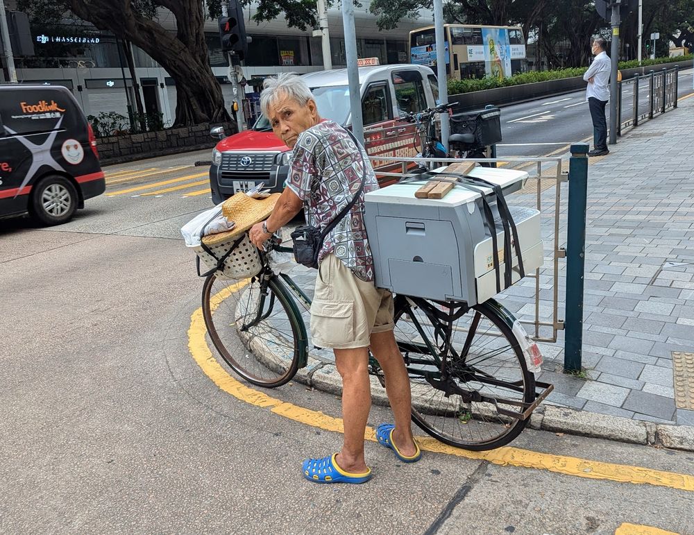 An older Chinese man in a patterned shirt and cargo shorts with blue and yellow crocs, walking a bike down the side of an urban street in Kowloon. There is a large office printer strapped to the back of the bike.
