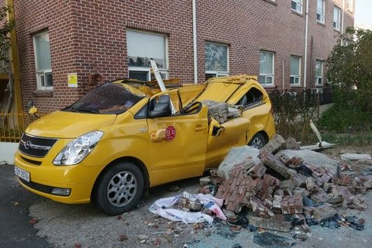 Yellow car damaged in 2017 magnitude 5.5 Pohang earthquake in South Korea. A pile of bricks sits next to the car, with a red brick building behind it. Credit: Sim1992/ Wikimedia Commons 
