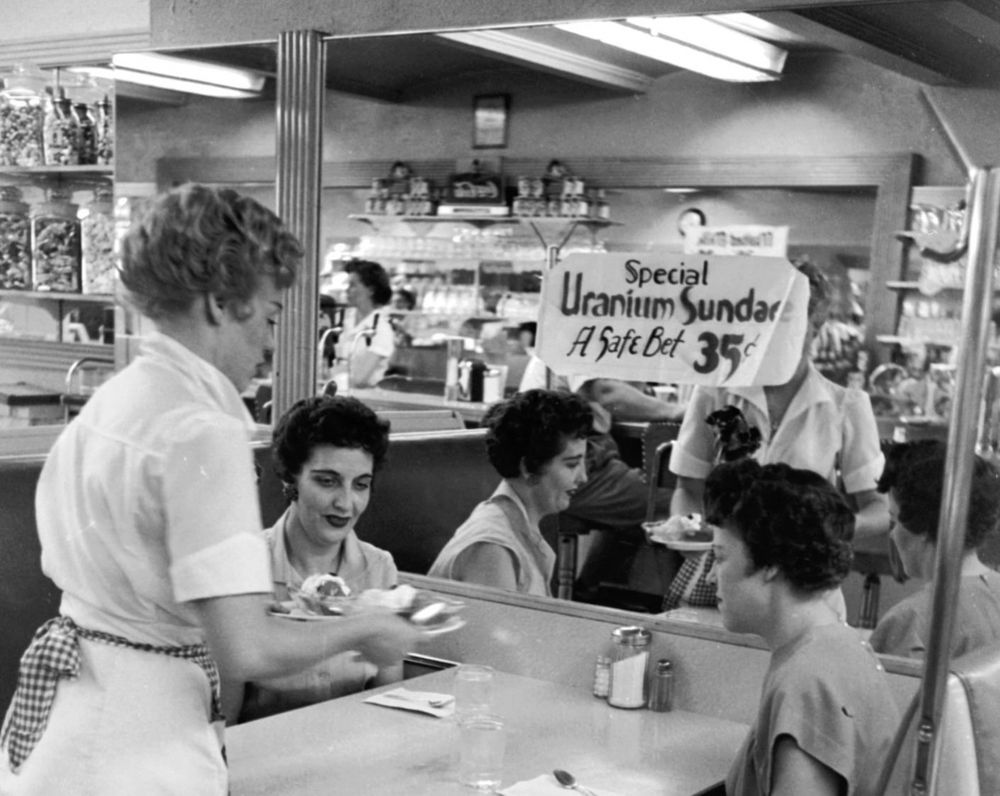 Black and white photo of a waitress serving a sundae in a cafe. A poster on the wall says “special: uranium sundae. A safe bet. 35c”