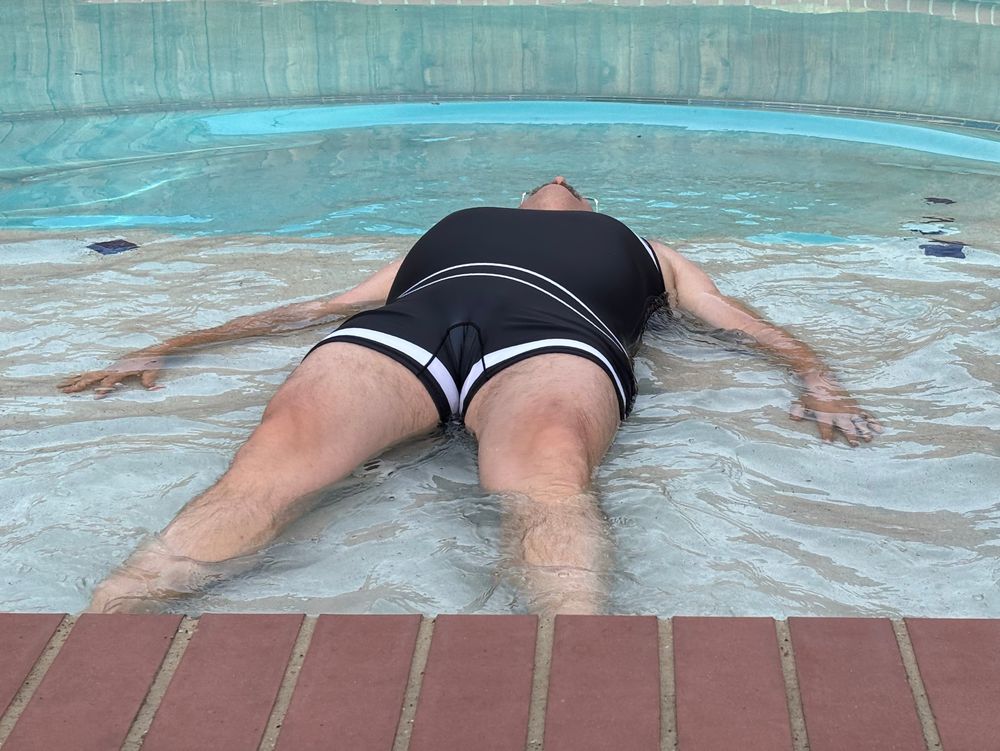 A guy in a black retro swimsuit laying on a shallow shelf in the pool