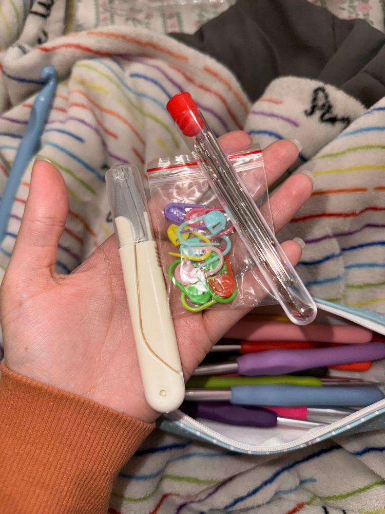 My hand, holding a pair of yarn scissors, a small bag of stitch markers, and a tube of blunt needles. In the background, there’s an open pouch filled with a bunch of ergonomic crochet hooks.