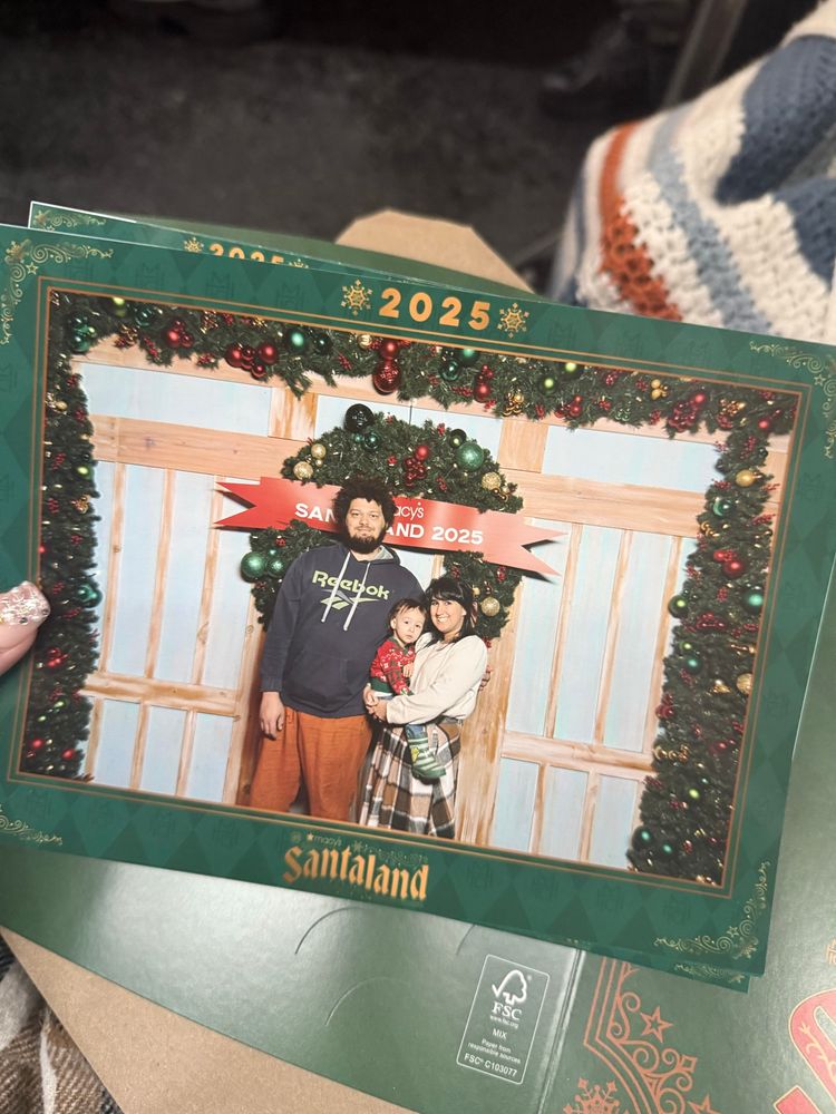 A printed copy of a family photo. The frame says 2025 on top and Santaland on the bottom. There is a very tall man blocking the sign that says Macy’s Santaland 2025. 