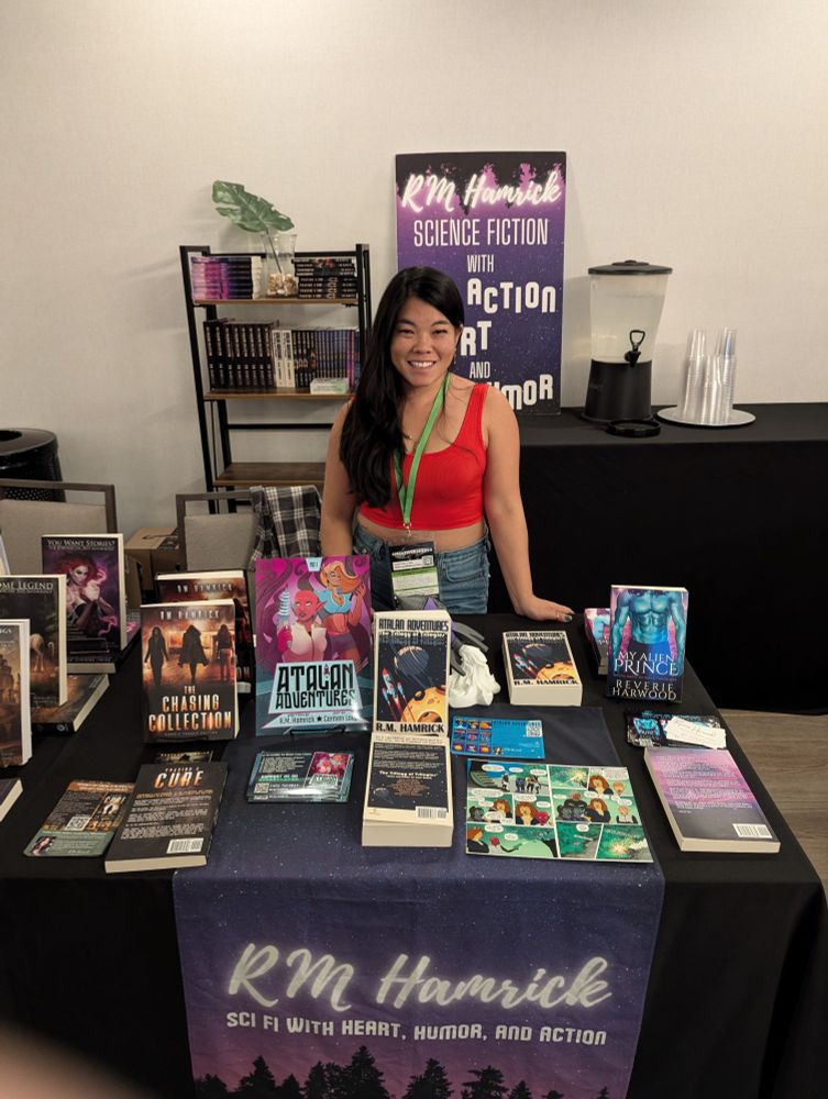 Woman with long black hair and red tank top smiling behind table of books she's written