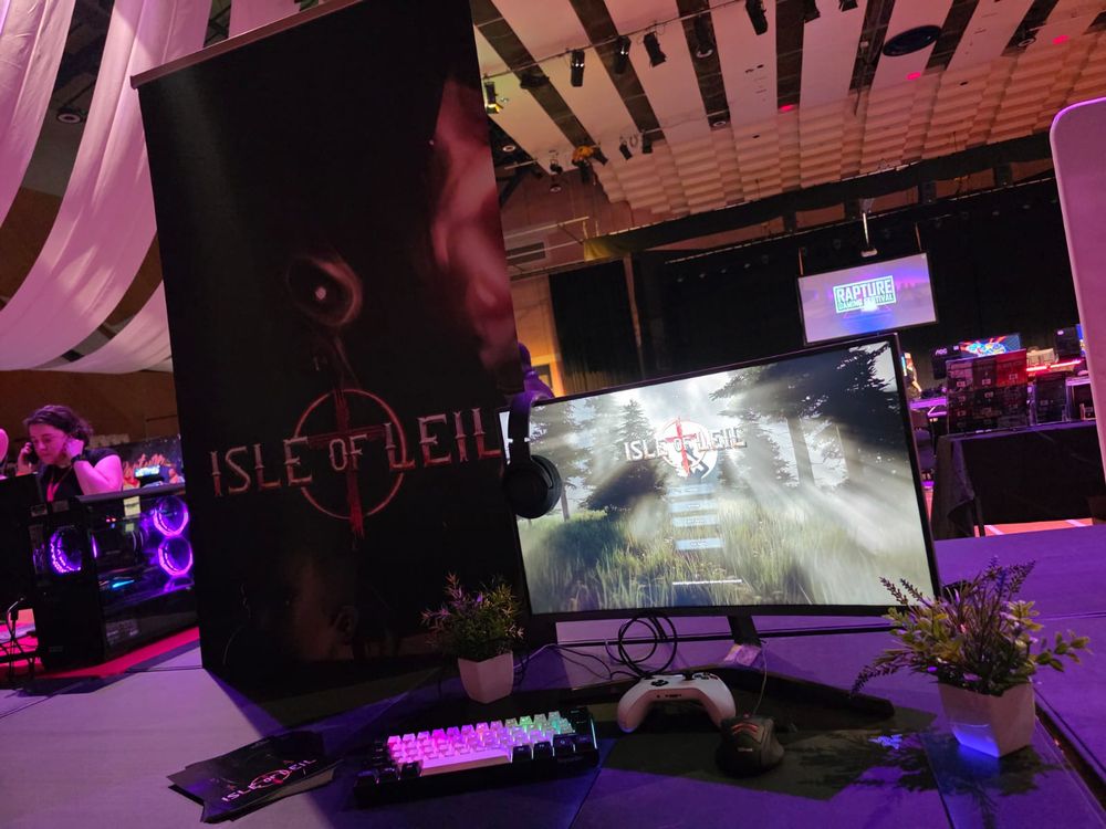 A photo of the set up for Isle of Leil at a gaming festival. A banner on the left with the logo of the game and a monitor to the right with the game loaded up, a controller and keyboard rest on the table in front.