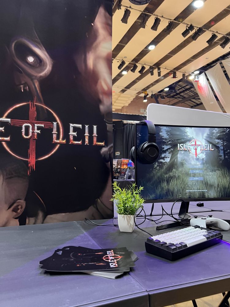 A photo of the set up for Isle of Leil at a gaming festival. A banner on the left with the logo of the game and a monitor to the right with the game loaded up, a controller and keyboard rest on the table in front.
