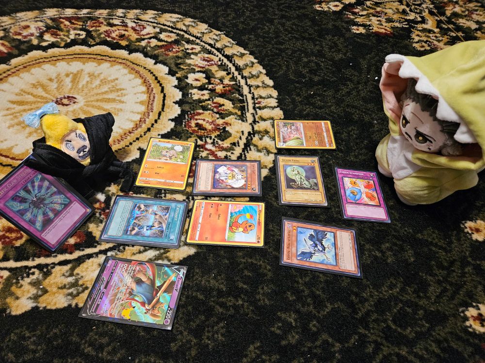 15 cm Izzy doll by imrights dressed in a dinosaur onesie and Star Izzy by disabledpirate dressed in a black robe. They have a spread of Pokémon and Yugioh cards between them and Star Izzy is holding a trap card from Yugioh
