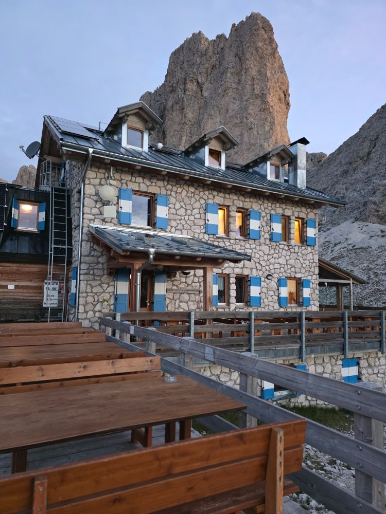 Eine Hütte aus hellgrauem Stein mit blau-weißen Fensterläden. Dahinter ein hoch aufragender Fels.
