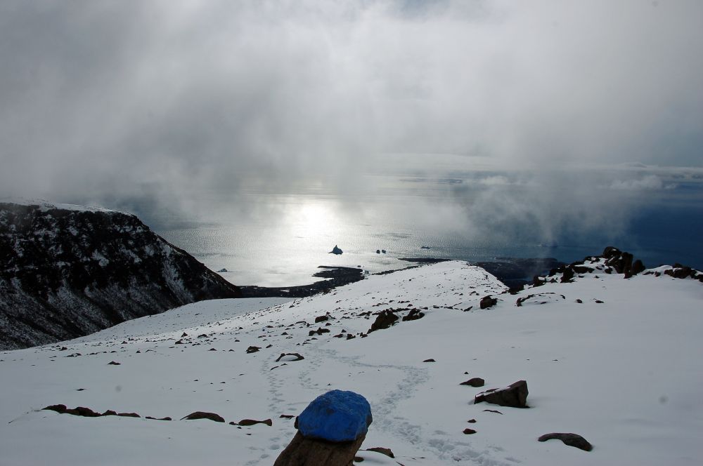 Blick von einem schneebedecktem Berg auf die Küste mit einem Eisberg. Der Blick ist wolkenverhangen. Im Vordergrund ein blau markierter Stein. 
