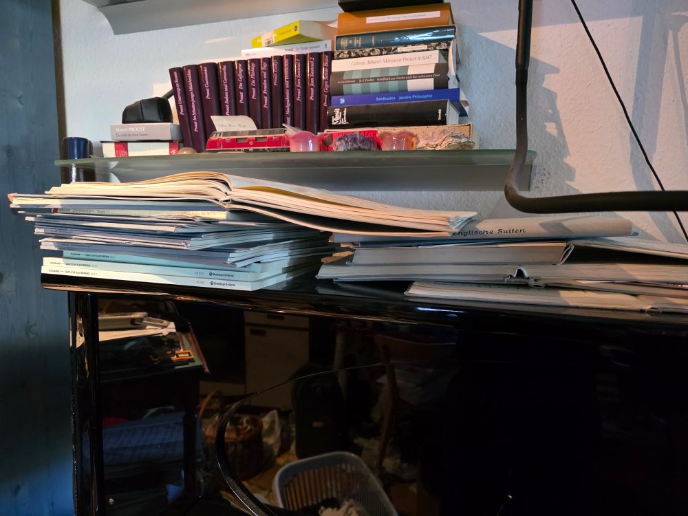 Ein schwarzes Klavier, darauf Stapel von Noten, teils aufgeschlagen, darüber ein Bücherregal. 