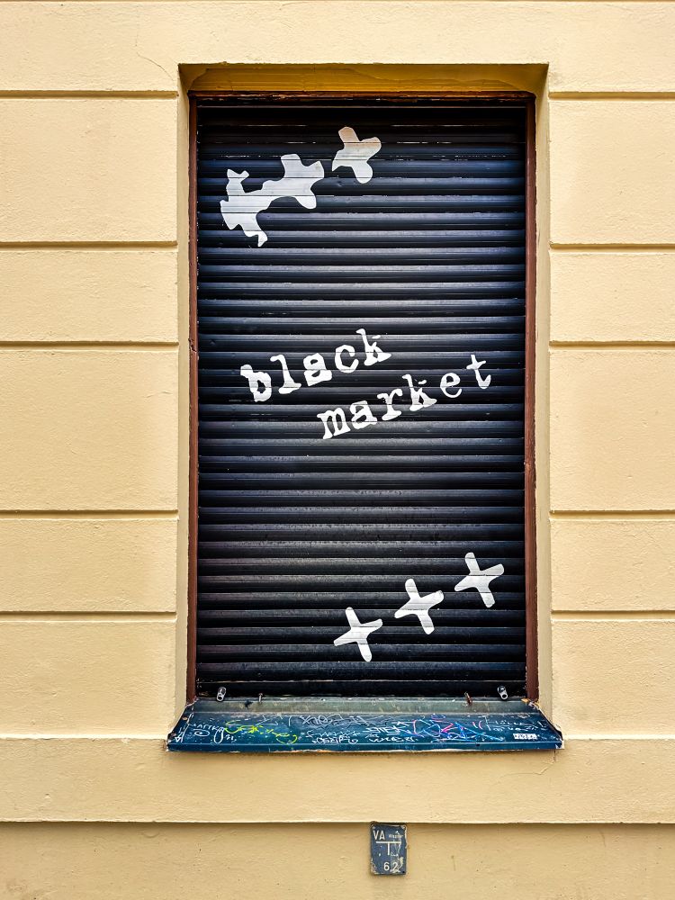 A shop window with a yellow-beige wall and a black metal shutter covering the window. On the shutter, the words “black market” are painted in white, uneven letters, set at an angle. Above and below the text are several white crosses, reminiscent of marks once used in telegrams to signify line breaks. The window ledge beneath the shutter is covered with colorful graffiti, adding to the urban character of the scene.