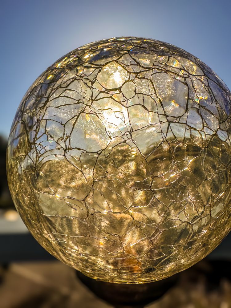 A  close-up view of a spherical glass object. The focus is sharp on the intricate, jagged network of cracks running throughout the surface, creating a mosaic-like appearance. Sunlight illuminates the glass, causing it to glow with warm golden hues that shimmer and reflect within the fractured patterns. The effect emphasizes both the delicate, almost crystalline structure of the glass and the vivid, radiant quality produced by the direct sunlight. The background is softly blurred, keeping all attention on the detailed, luminous texture of the glass sphere.