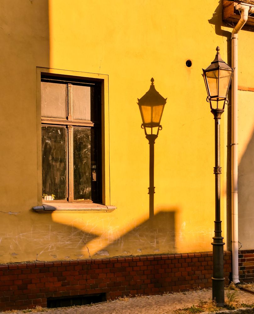 The image shows a yellow house wall with a window on the left side and a classic street lamp on the right. The sun is low, casting a sharp shadow of the street lamp onto the wall. The lower part of the wall features red bricks. The scene is bathed in warm, golden sunlight, creating long shadows and a calm atmosphere.