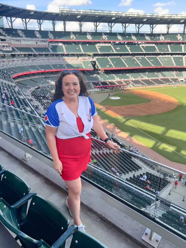 A brunette woman wearing a short red dress with a cropped blue and white Braves city connect Jersey stands on a partially sunny day overlooking Truist Park
