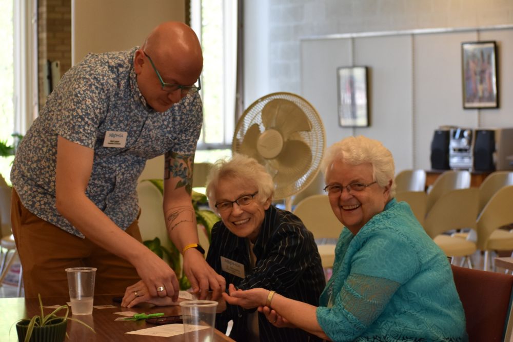 A bald man with glasses in a short sleeve floral print shirt and khaki pants putting fake tattoos on two women, one in a teal shirt and one in a darker shirt, both have shirt white hair and glasses and are looking at the camera and giggling because they think it's kind of ridiculous and silly that they are getting fake tattoos. But they are also really enjoying themselves. 