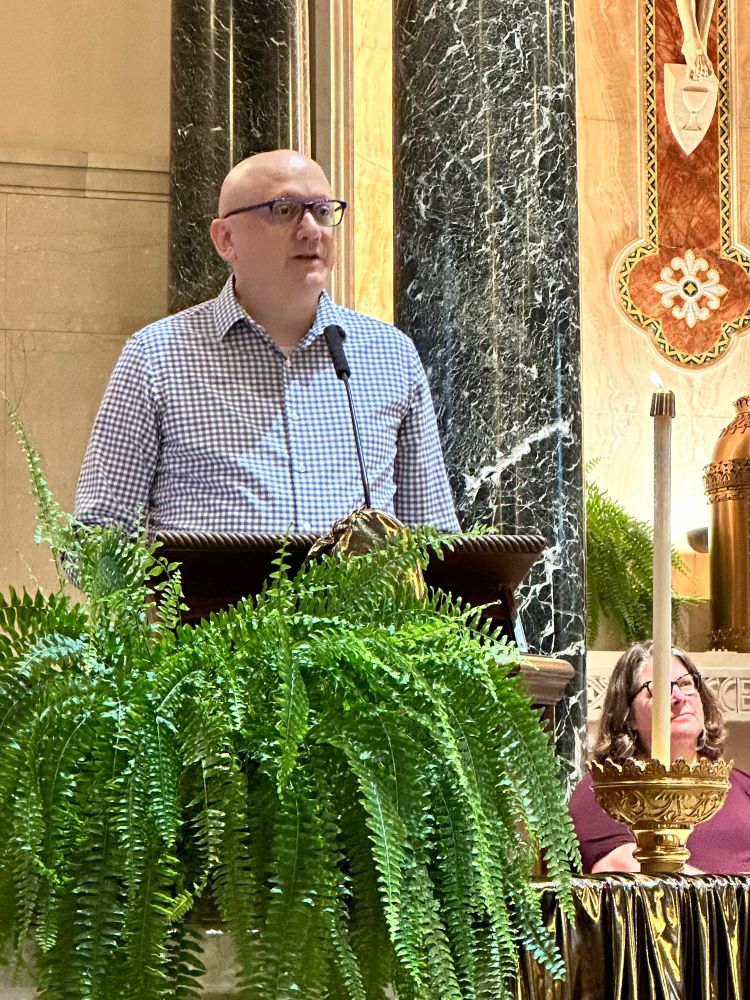 Here I am sharing my own personal definition of spiritual direction with the assembled peoples. I'm a bald white guy with glasses wearing a gray and white gingham checkered shirt. There's a large fern in front of the lectern where I am speaking. There's a candle to my left and you can kind of see my friend Heather sitting behind the candle. 