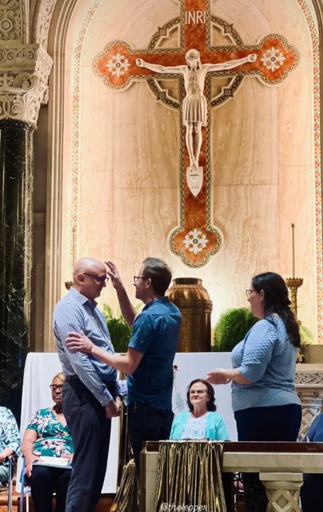 A bald man in a long sleeved white and gray gingham button up shirt and gray pants facing another man in a short sleeved blue shirt. The bald man is being anointed with oil by the other man. Behind them are several women sitting in chairs and directly behind and do the left of the main doing the anointing is a woman with long brown hair in a light blue shirt holding the dish of oil. On the wall in the background is a beige and red marble crucifix. 
