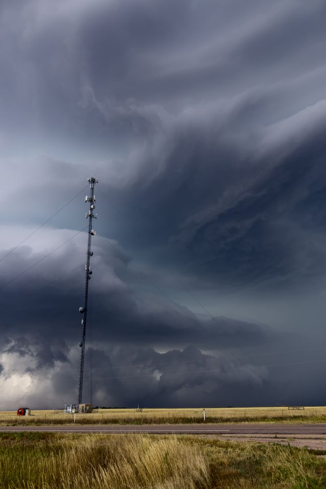 A road passes through the bottom of the image with dead grass visibly above and below it. Small structures are seen in the distance, with a large radio tower with several ground-attached wires strung throughout the entire pole. A massive storm appears in the back with distinct shapes - a ragged tail cloud fills into the storm on the bottom third of the image, with the main storm base above it and to the left. Small, ragged clouds decorate the rest of the underside of the base. Above the base extends a greater storm plateau- with an aesthetic akin to ocean waves as the plate of clouds extends and lightens upwards. This plate takes up the upper half of the image, with darker portions visibly more turbulent.