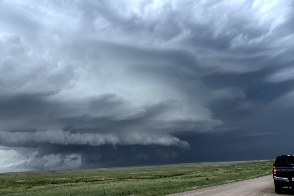 Soft green grass covers the bottom fifth of the image with a pale dirt road cutting through the far right. Sitting on that road is a black truck, only partially visible.  large storm takes up the majority of the frame with a body that looks like sculpted pottery (smooth, wavy lines). It's ornamented with a raggedy, short horseshoe shaped shelf cloud. This photo was taken on June 29th, 2023 and photographs a supercell west of Last Chance, Colroado.