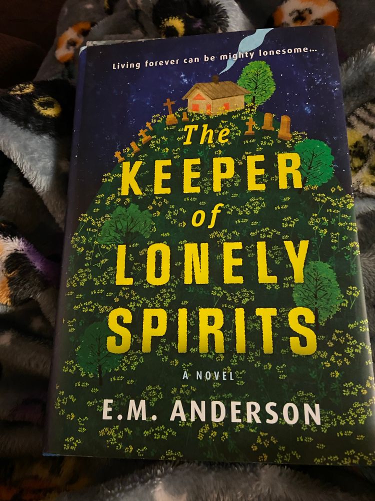 The cover of The Keeper of Lonely Spirits by EM Anderson. It shows a house, surrounded by graves, on a hill