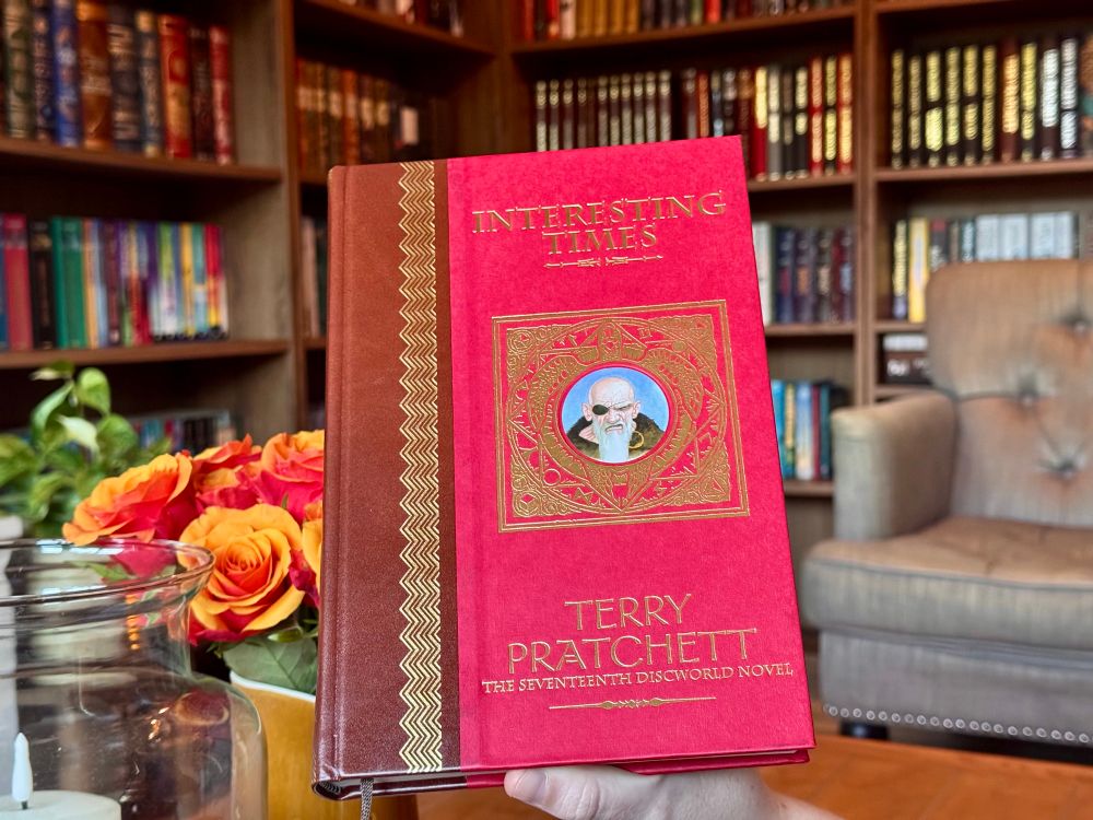A red leather-bound book (Interesting Times by Terry Pratchett) in front of red roses and bookshelves.