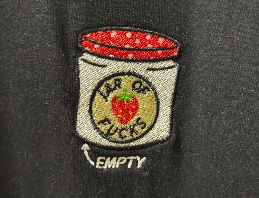 A grey t-shirt with an embroidered jam jar on it. One can read jar of fucks on it. An arrow below reveals it's empty.