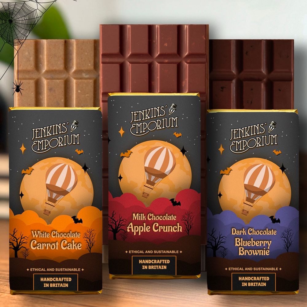 Three chocolate bars popping from behind their wrappers: White chocolate carrot cake, Milk chocolate apple crunch, and dark chocolate blueberry brownie. The wrappers are showing a hot air balloon flying in front of a yellow moon under a black sky. Bats are flying here and there.