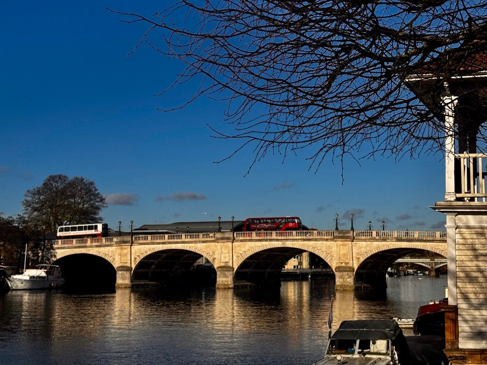 A stone bridge over the Thames. A red bus crossing it. Blue skies up.