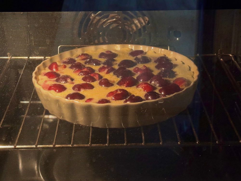 A ceramic pie dish in an oven. Red cherries are floating in batter inside it.