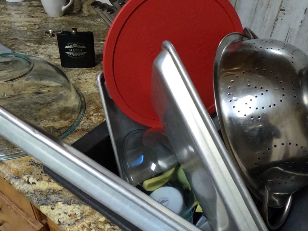 A pile of kitchen stuff drying in a sink after being handwashed: Metal trays, big red mixing bowls, a colander....an open flask is visible at rear.