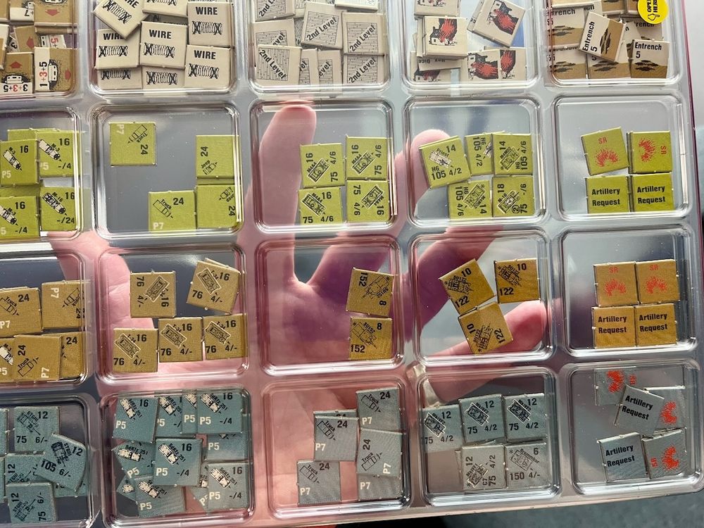 A hand holds up a translucent plastic tray whose compartments are filled with green, tan and blue cardboard counters from the Avalon Hill wargame, Squad Leader.
